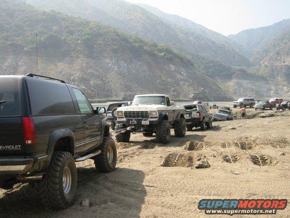 Several lifted trucks and SUVs lined up on a dirt road with mountains in the background. One truck has a white cab and brown bed
