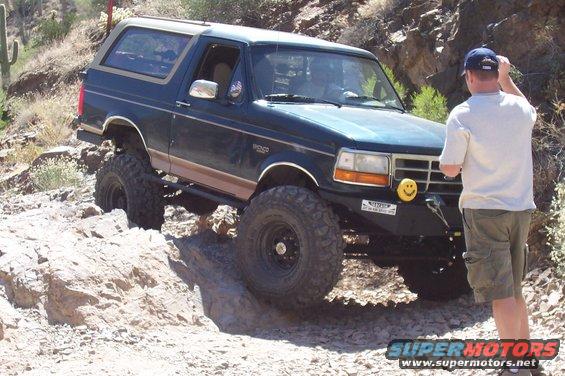 Dark blue Ford Bronco with lifted suspension and large tires navigating a rocky trail. A man stands nearby