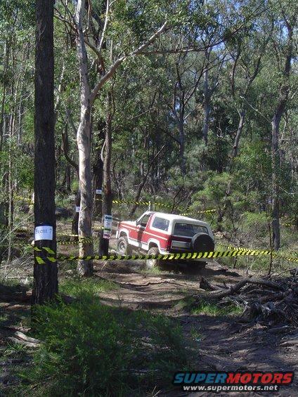 Red and white SUV navigating a dirt path in a wooded area, with yellow caution tape marking a course