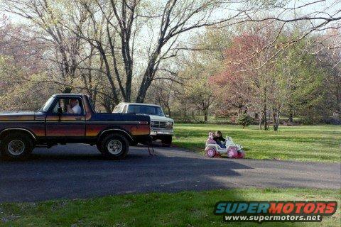 Ford Bronco parked next to a white SUV, with a child in a pink toy car driving past