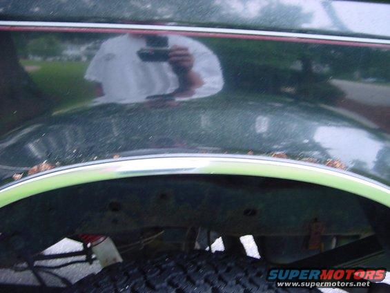 Rear fender wheel well and tire of a dark green vehicle, with chrome trim. Reflection of a person holding a camera is visible