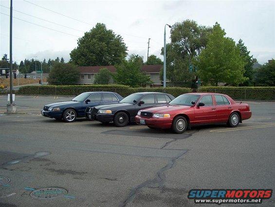 Three police sedans parked in a lot. One is red, two are dark blue/black