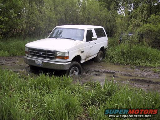 White 1992 Ford Bronco stuck in mud, front 3/4 view, grass and trees surrounding