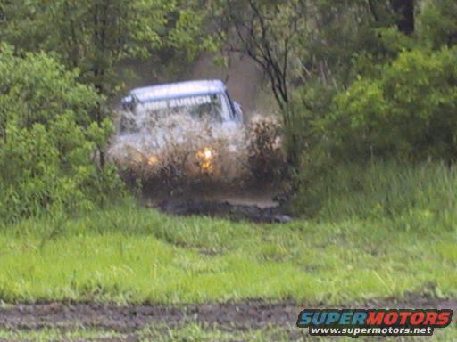 Off-road vehicle driving through a muddy puddle, splashing water. Text on vehicle reads 'ZURICH'