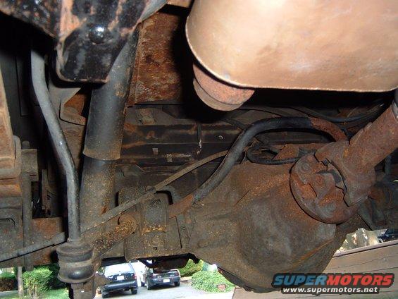 Underside view of a rusty rear axle, driveshaft, and suspension parts. Two cars are visible in the background