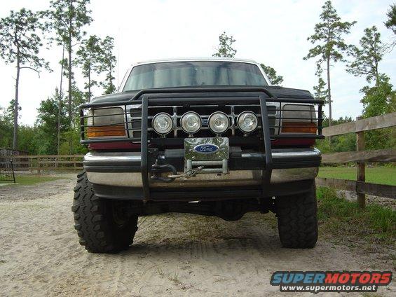 Front view of a dark colored Ford truck with a black bull bar, four auxiliary lights, and a winch. It has large off-road tires