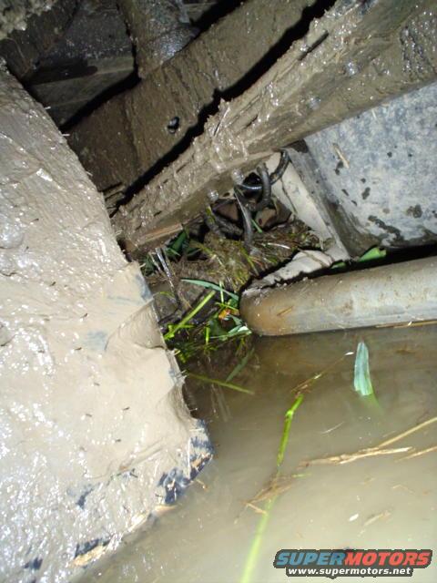 Mud-covered vehicle frame and suspension submerged in murky water with grass