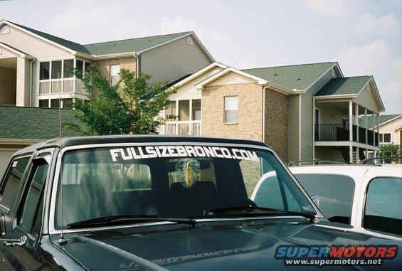 Dark gray Ford Bronco with a "FULLSIZEBRONCO.COM" sticker on the windshield parked next to a white SUV. Apartment buildings in the background