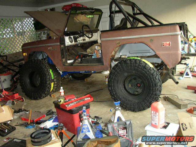 Brown 1974 Ford Bronco with large off-road tires, roll cage, and hood open, in a garage