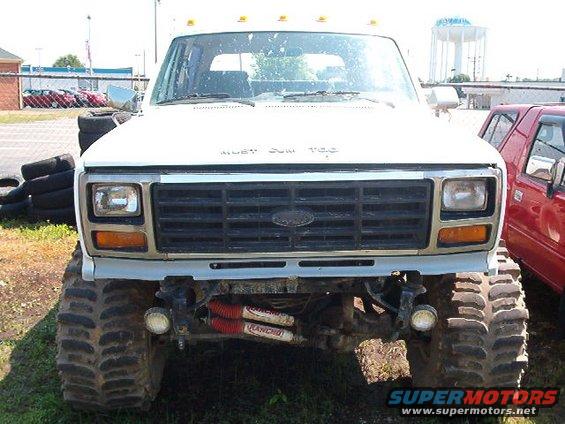 Front view of a lifted Ford F-Series truck with large off-road tires. A red vehicle is partially visible to the right