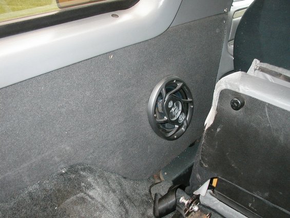 Aftermarket speaker mounted on grey carpeted panel inside a vehicle, next to a black seat