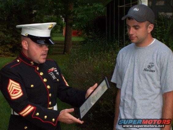 Marine in dress blues presents a framed document to a man in a gray t-shirt with a Marines logo