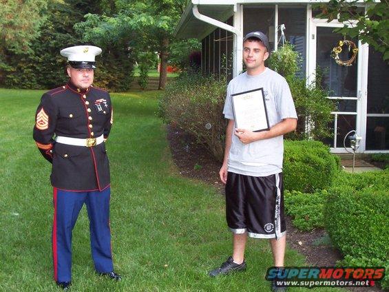 Man in Marine dress uniform stands next to a man holding a framed document