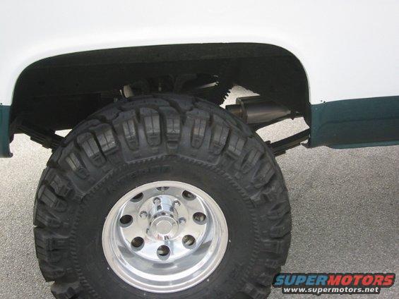 Close-up of a large off-road tire and chrome rim on a lifted truck, showing leaf springs and exhaust pipe