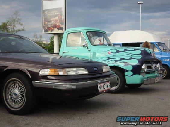 Dark sedan parked next to a teal pickup with blue flames. A blue van is behind the truck