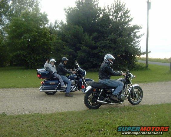 Two motorcycles are ridden on a gravel path. The lead motorcycle has one rider. The second motorcycle has two riders, with the passenger giving a peace sign
