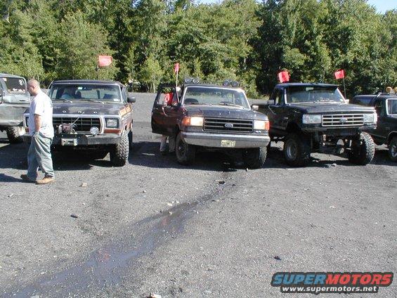 Three Ford trucks parked in a gravel lot. The leftmost truck is a dark color with a winch. The middle truck has its door open. The rightmost truck is lifted