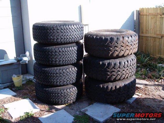 Stack of six off-road tires, some with aggressive tread patterns, outdoors near a wooden fence