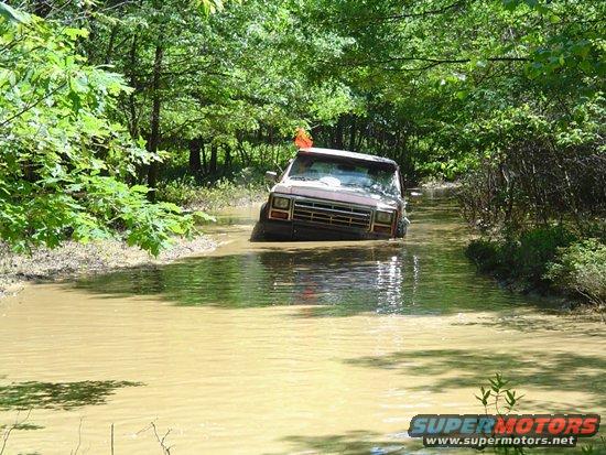 Brown SUV driving through a muddy water-filled path in a wooded area. Water is up to the doors