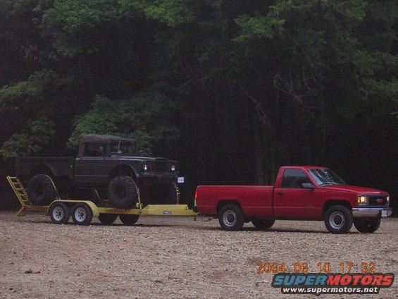 Dark off-road truck on yellow trailer towed by red GMC pickup, side view