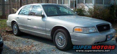 Silver 2000 Ford Crown Victoria parked on gravel with trees and a building in the background