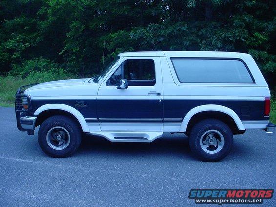 Side view of a white and dark blue 1995 Ford Bronco parked on asphalt with trees in the background