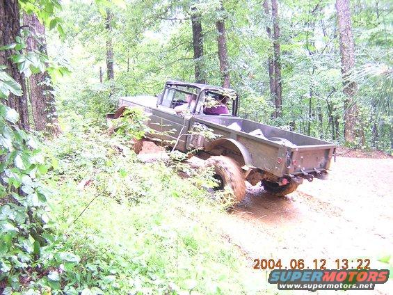 Olive green off-road truck with open bed driving through mud on a wooded trail