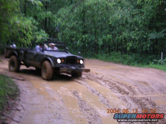 Dark green Jeep Gladiator J-series pickup truck driving through mud, front-left view. Visible roll bar, open top, and off-road tires