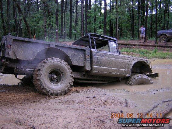 Muddy off-road truck stuck in a mud pit with trees in background. Another vehicle visible to the right