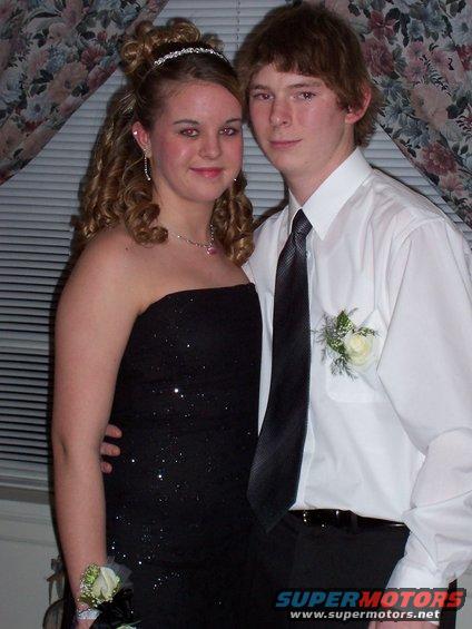 Teen couple dressed up for a formal event. Girl wears a sparkly black dress and tiara. Boy wears a white shirt and tie with a boutonniere