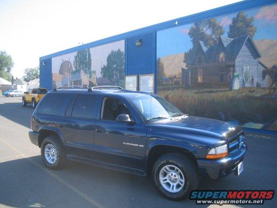Dark blue SUV parked in front of a building with a mural. Another yellow truck is visible in the background