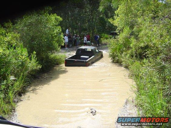 Black pickup truck partially submerged in muddy water with people watching on the bank