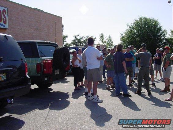 Green Ford Bronco with spare tire on back, parked next to a dark SUV, with a group of people standing around