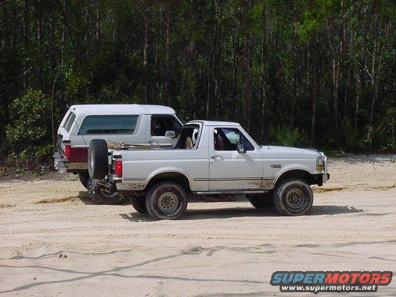 Two white Ford Broncos parked on a dirt road, one behind the other. The front Bronco is a convertible with the top removed