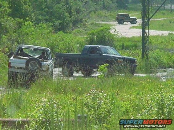 Two off-road vehicles are driving through a muddy area with trees and grass around. Another vehicle is visible in the background