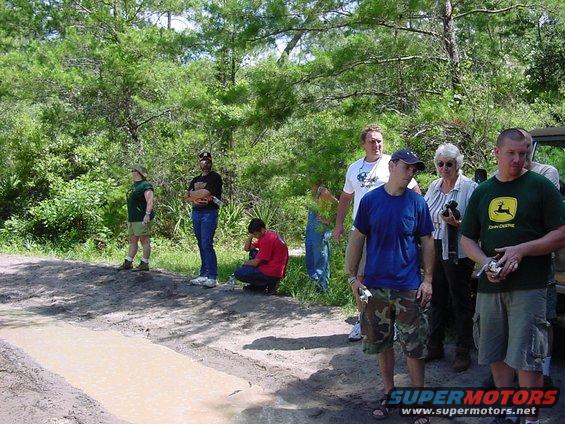 People gathered on a dirt path next to a muddy water obstacle