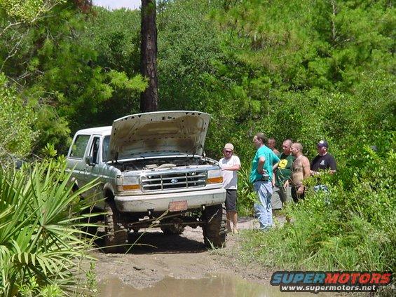 Silver Ford Bronco with hood open stuck in mud, seen from front-right