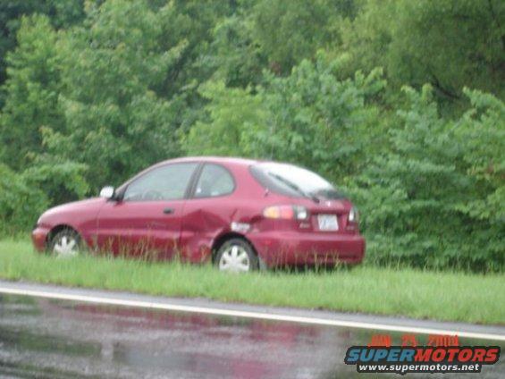 Red hatchback car with damage to rear quarter panel, parked on grassy shoulder next to a road