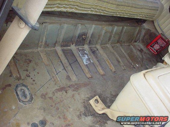 Interior floor of a vehicle with visible rust and metal ribs, a red book, and beige seats
