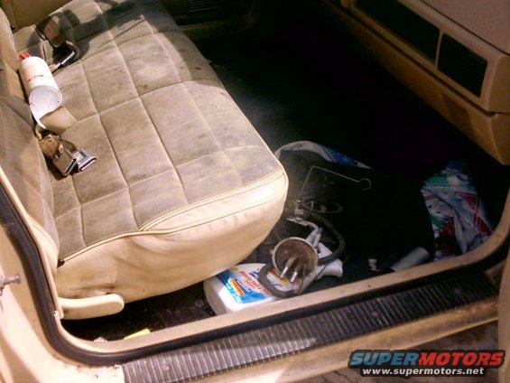 Interior of a vehicle showing a dirty, quilted seat and cluttered floor with a bottle of cleaner and car parts