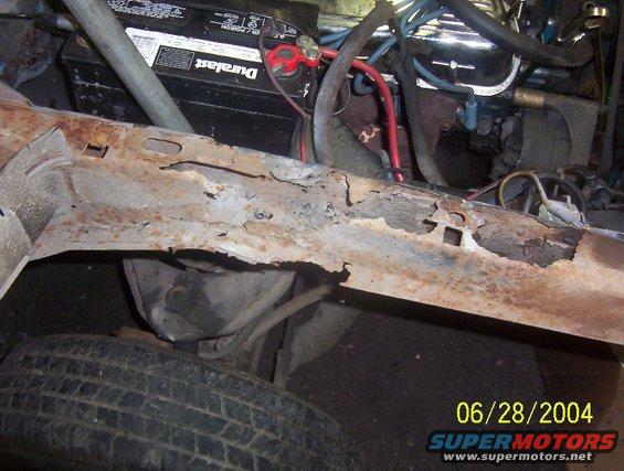 Rusted vehicle frame section with battery and engine parts visible, tire in foreground