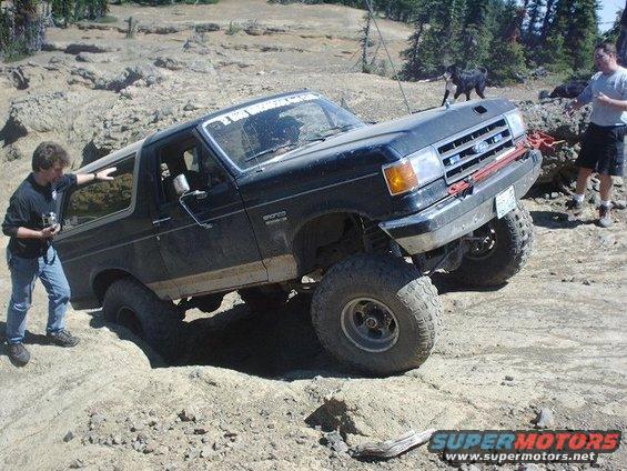 Black Ford Bronco with lifted suspension navigating a rocky trail. A person stands by the driver's door, another person and a dog are in the background