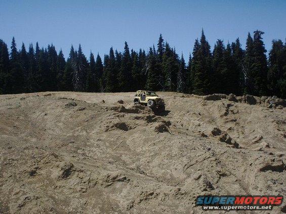 Yellow off-road vehicle climbing a dirt hill with pine trees in the background. Watermark reads SUPERMOTORS