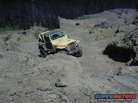 Yellow Jeep CJ-7 navigating a steep, rocky trail with trees in the background