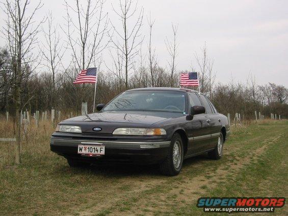 Dark 1992 Ford Crown Victoria sedan with two American flags on poles, parked on a grassy area