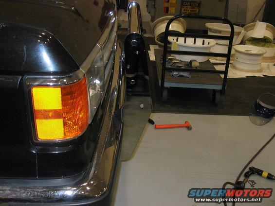 Black SUV rear corner with chrome bumper and amber turn signal. A cart with white rims is nearby