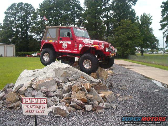Red Jeep Wrangler with "SkyJacker" decals, lifted on large tires, perched on a pile of rocks