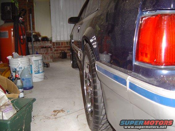 Dark blue car, likely a 1987 Ford Mustang, parked in a garage. Visible details include the rear tail light, side trim, and wheel