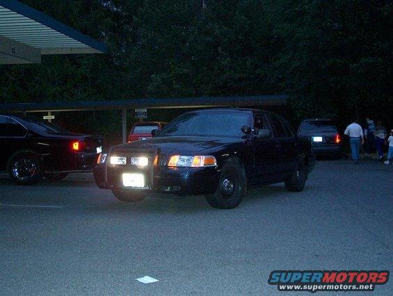 2001 Ford Crown Victoria police car parked, front view. Another car is to the left, and people are gathered in the background