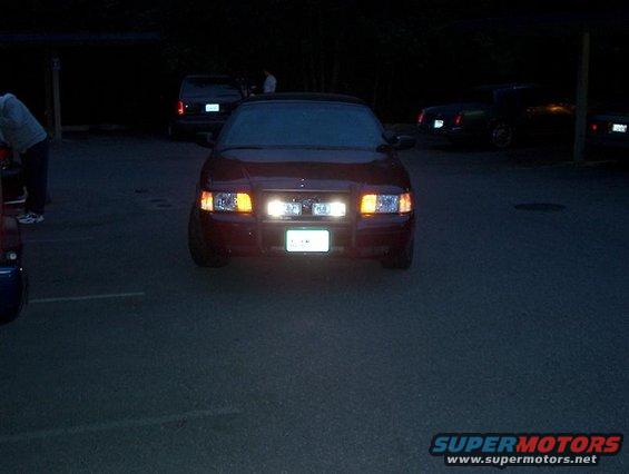 Front view of a red sedan with headlights on. Other cars are parked in the background